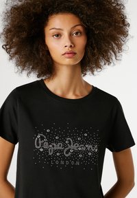 Mladá žena s kučeravými vlasmi, oblečená v čiernom tričku so striebornými nitmi a vzormi na jednoduchom bielom pozadí.