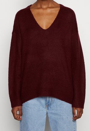 Henkilö, jolla on päällään löysä, tumman viininpunainen V-kaula-aukollinen neule, jossa on resorit hihansuissa, ja vaaleansiniset farkut, kuvattuna olkapäistä lantioihin.