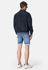 Granatowa bomberka z prążkowanymi mankietami i kołnierzem, zestawiona z jasnymi krótkimi spodniami dżinsowymi, widocznymi szwami oraz białymi trampkami. Gładka faktura materiału.