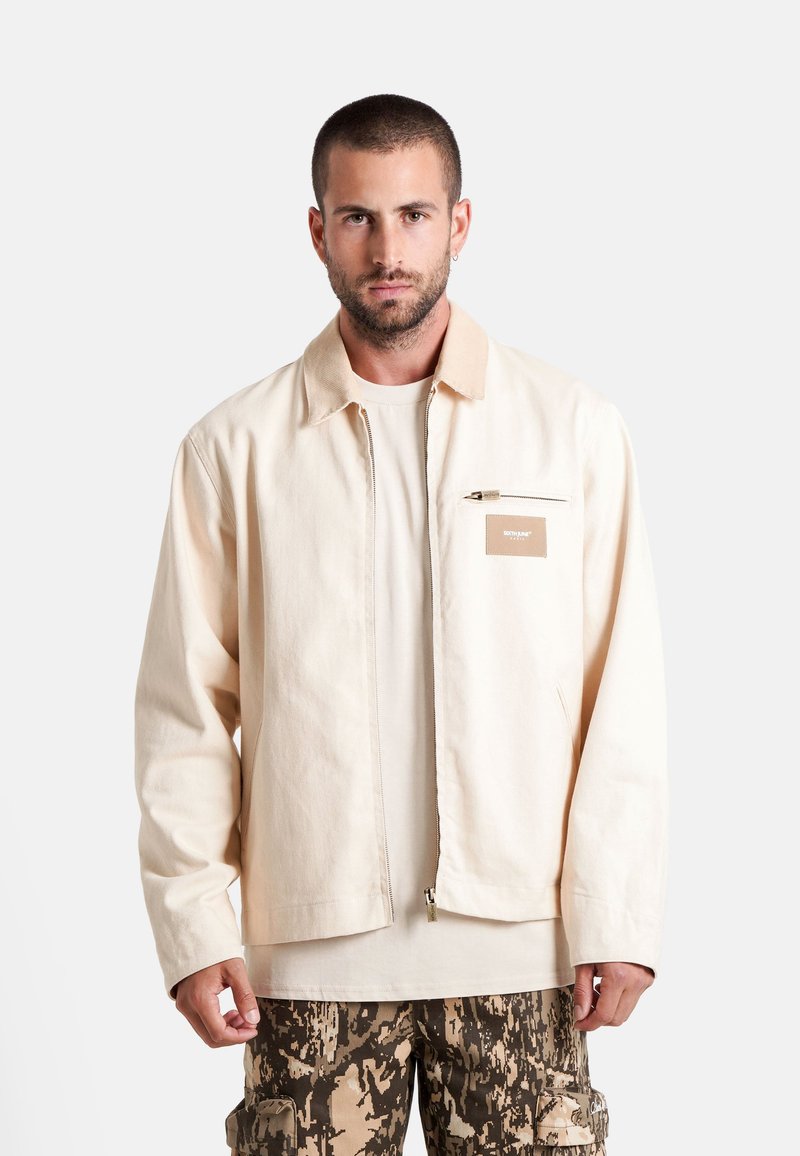 Lichtbeige canvas jack met een kraag, voorzichtige rits en een borstzak. Draag het over een crèmekleurig shirt en gestreepte cargobroek.