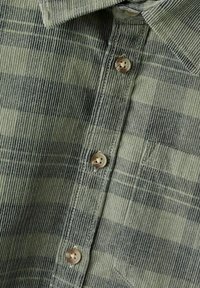 Grønn og grå rutete kordfløyelsskjorte med krage, med knappelukking og teksturert stoff. Lett broderte detaljer er synlige.