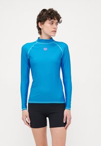 Camisola de manga longa em azul brilhante, feita de tecido elástico. Apresenta costuras em contraste e um logo rosa no peito. Combinada com calções pretos.