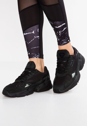 Person wearing black Adidas sneakers with mesh and suede details, paired with black leggings featuring white marble-patterned panels.