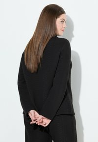 Kobieta z długimi brązowymi włosami, ubrana w czarną, teksturowaną bluzkę z długim rękawem i spodnie, stoi z rękami złożonymi za plecami, zwrócona w prawo.