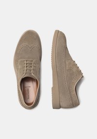 Chaussures en suede beige avec des détails brogue, à lacets et avec une semelle en caoutchouc texturée. Elles présentent un embout rond et un intérieur lisse.