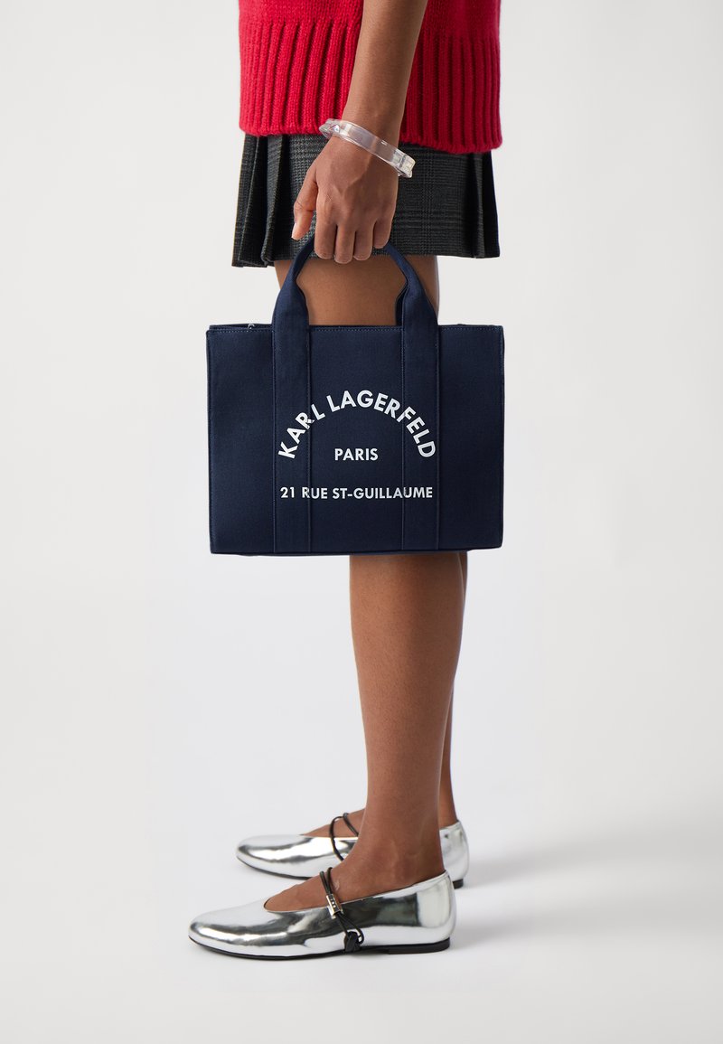 Navy blue tote bag with "Karl Lagerfeld Paris" and address in white text, featuring sturdy handles. Model wears silver ballet flats.