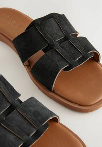 Svarta mocka slide-sandaler med två breda remmar, med ett rektangulärt mönster och bruna gummisulor. Framträdande sömnadsdetaljer längs kanterna.