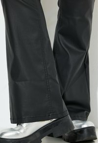 Zwarte, waterdichte broek met een gladde textuur, uitlopende snit en zichtbare stiksels. Gecombineerd met glanzende zilveren enkellaarsjes en dikke zolen.