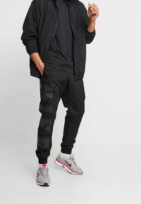 Man som bär svart träningsjacka och byxor med stor grafisk text på vänster ben, vita strumpor och grå och röda Nike-sneakers.