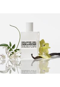 Heldere fles Zadig & Voltaire parfum met een witte dop, zwart gelabeld. Omringd door witte bloemen en groene bladeren, op een reflecterend oppervlak.