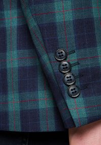 Wełniana marynarka w granatowo-zieloną krateczkę z czerwonymi akcentami. Posiada cztery czarne guziki oraz wyraźną fakturę z tkaniny w tkanym wzorze.