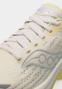 Κοντινό πλάνο ενός παπουτσιού για τρέξιμο Saucony σε κρεμ και ανοιχτό κίτρινο χρώμα, με λευκά κορδόνια και διαπνέον υφασμάτινο πλέγμα.