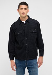 Schwarzes Cordhemd mit langen Ärmeln, zwei Brusttaschen und Knopfleiste. Darüber ein weißes T-Shirt getragen, kombiniert mit blauen Jeans.