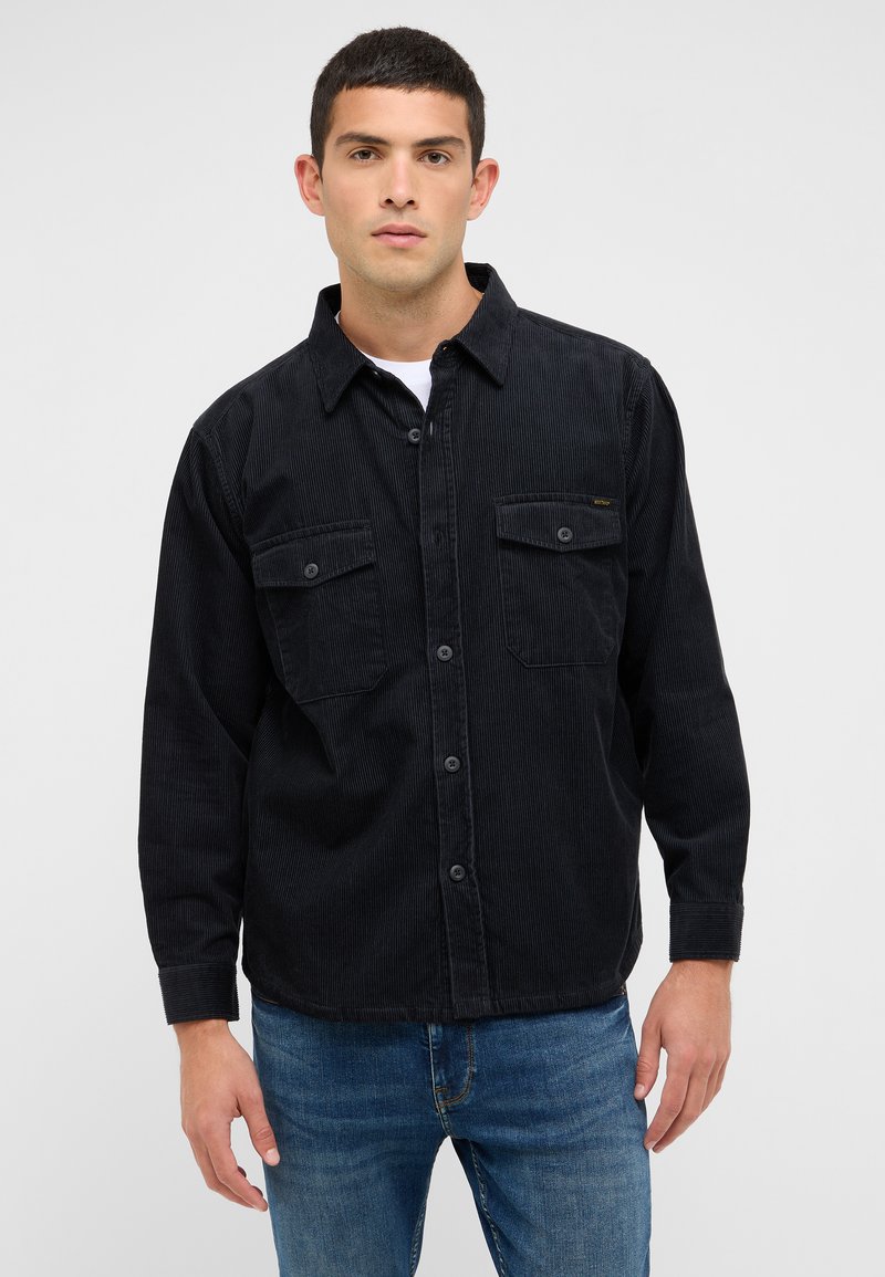 Schwarzes Cordhemd mit langen Ärmeln, zwei Brusttaschen und Knopfleiste. Darüber ein weißes T-Shirt getragen, kombiniert mit blauen Jeans.