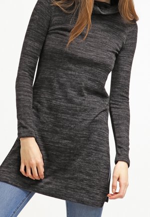 Vrouw draagt een donkergrijze tuniek met lange mouwen en een lichte zijsplit, gecombineerd met blauwe jeans, tegen een witte achtergrond.