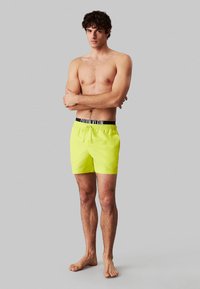 Neon gele zwembroek gemaakt van lichtgewicht stof, met een elastische zwarte tailleband met witte branding en een trekkoord voor aanpassing.