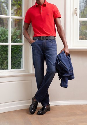Rotes Poloshirt, dunkelblaue Jeans, schwarzer Gürtel und glänzende schwarze Loafers. Hält eine dunkelblaue Jacke, steht an einem Fenster mit Regen.