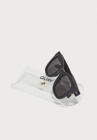 Lunettes de soleil noires surdimensionnées avec une monture géométrique et des verres foncés, placées dans un étui de protection transparent avec une fermeture à bouton doré.