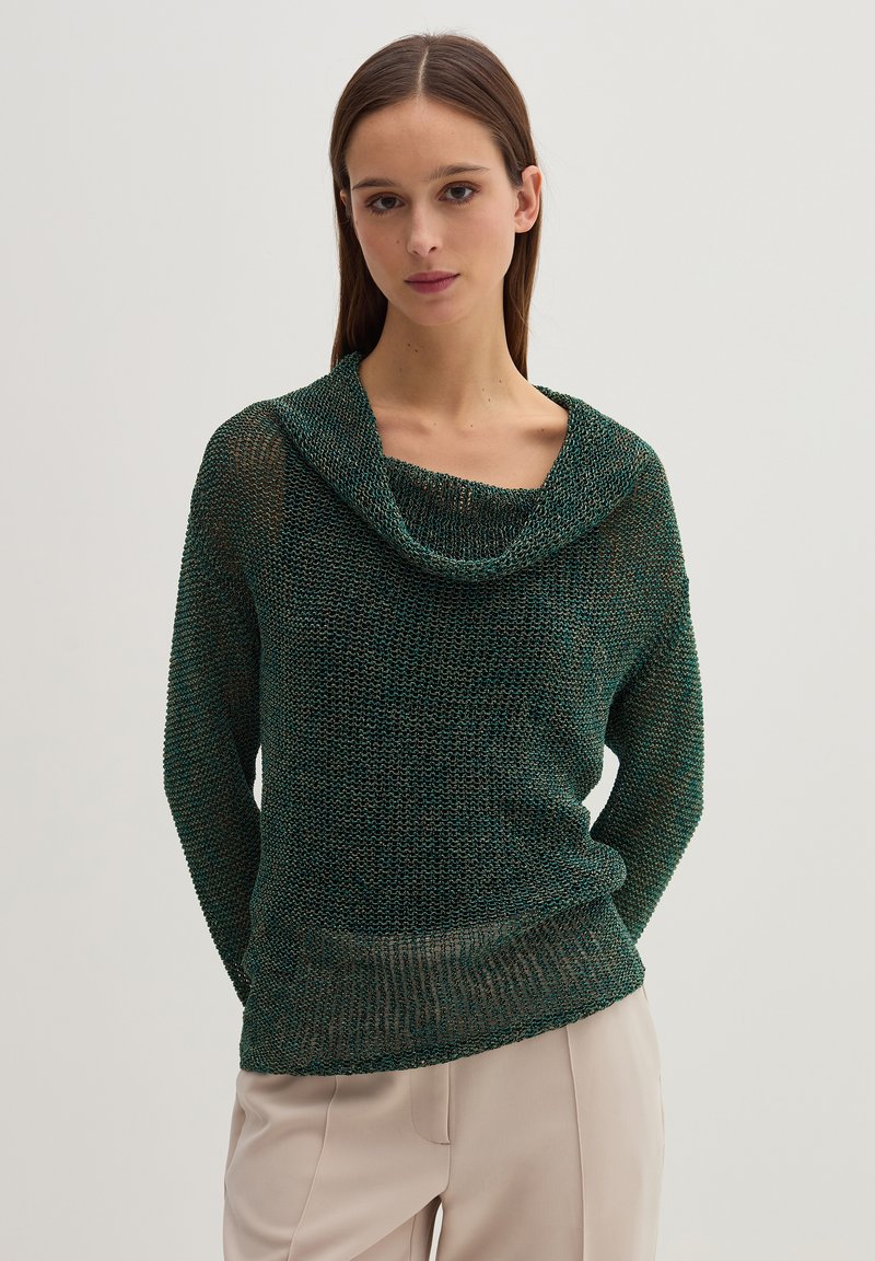 Suéter verde de punto con cuello de cisne, con un corte holgado y tejido texturizado. Combinado con pantalones de color claro.