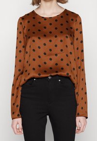 Femme portant un chemisier marron à manches longues avec des pois noirs, rentré dans un pantalon noir taille haute, sur un fond uni.