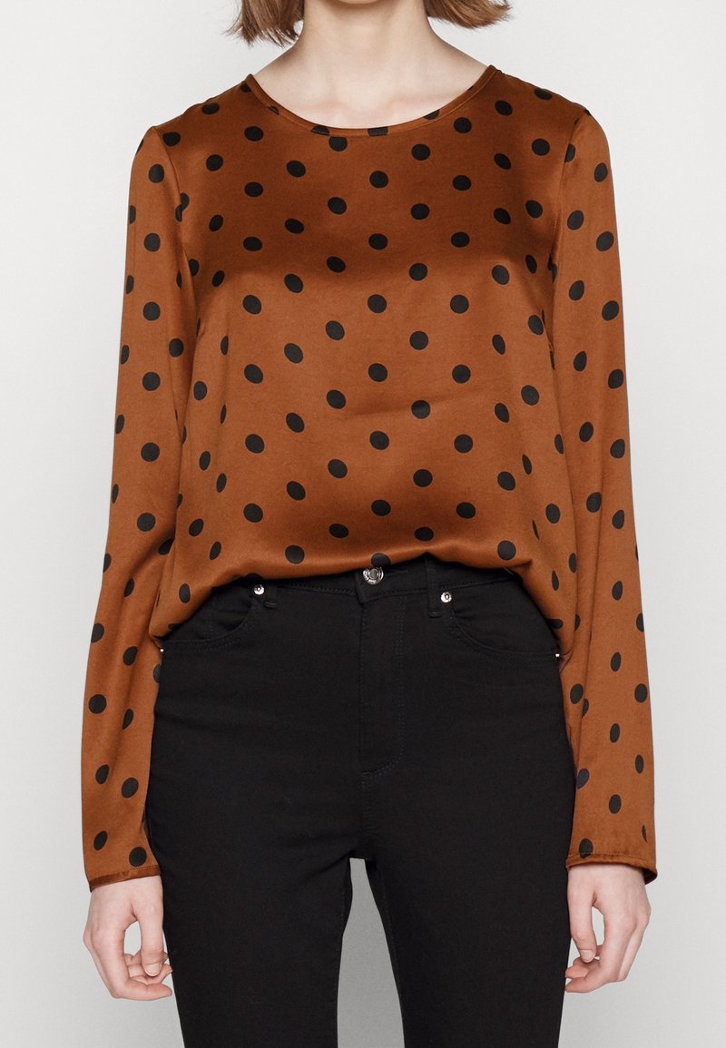 Femme portant un chemisier marron à manches longues avec des pois noirs, rentré dans un pantalon noir taille haute, sur un fond uni.