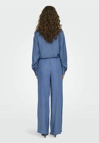 Woman with long curly hair wearing a blue long-sleeve jumpsuit and heels, standing facing away against a plain background.