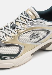Sneaker Lacoste z białą i złotą siateczkową cholewką, szarymi akcentami i sznurowadłami, teksturowaną podeszwą oraz logo z boku.