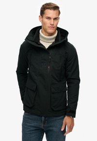 Schwarze wasserdichte Jacke mit Kapuze, zwei Fronttaschen, einer mit Reißverschluss versehenen Brusttasche und strukturiertem Stoff. Getragen über einem beigen Pullover.