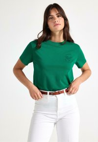 Grøn bomulds t-shirt med rund hals og korte ærmer, med et sort trykt tennislogo på brystet. Båret sammen med hvide højtaljede bukser.