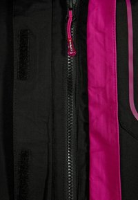 Giacca nera con un rivestimento interno rosa a contrasto, una zip prominente con un tirante rosso e una patta in velcro sul lato sinistro.