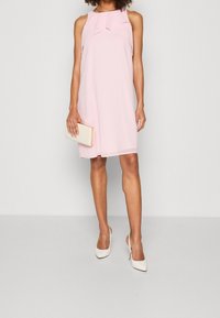 Donna che indossa un vestito senza maniche di un rosa pallido, lungo fino al ginocchio, con dettagli a volant, che tiene una pochette beige e indossa scarpe con tacco alto bianche.