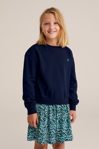 Mornarsko modra jopica s rebrastimi manšetami in robom, v kombinaciji z zeleno obleko s cvetličnim vzorcem, ki vsebuje pliseje in elastičen pas.