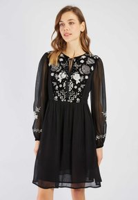 Zwarte jurk met doorschijnende lange mouwen, geborduurde bloemen- en geometrische patronen op het lijfje, trekkoord bij de hals en een vloeiende rok.