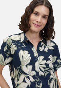 Marinebluse mit kurzen Ärmeln, die große weiße und grüne Blumenmuster aufweist, Knopfleiste vorne und einen spitzen Kragen hat. Glatte Stofftextur.