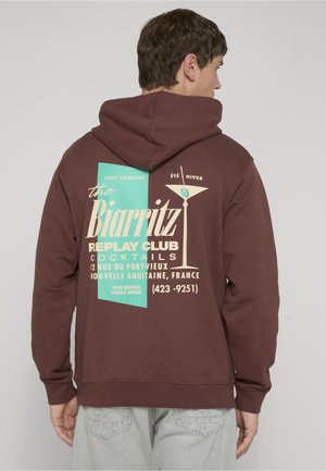 Mężczyzna ubrany w brązową bluzę z kapturem z turkusowo-beżowym nadrukiem Biarritz cocktail club oraz napisem lokalizacji na plecach, zwrócony tyłem do kamery.
