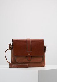 Brązowa skórzana torba typu crossbody Fossil z przednią klapą, magnetycznym zapięciem i regulowanym paskiem na białym piedestale.
