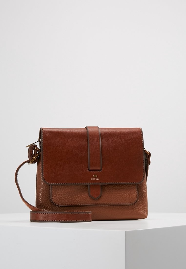 Brązowa skórzana torba typu crossbody Fossil z przednią klapą, magnetycznym zapięciem i regulowanym paskiem na białym piedestale.
