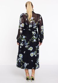 Schwarzes, durchsichtiges Maxikleid mit floralem Muster in Blau und Weiß, tailliert mit Gürtel, langen Ärmeln und fließender Silhouette. Sichtbare grüne Stiletto-Absätze.