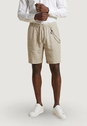 Mężczyzna ubrany w beżowe lniane szorty ze ściągaczem i ozdobą w postaci łańcucha, białą koszulę z długim rękawem oraz białe trampki, stoi ze splecionymi rękami.