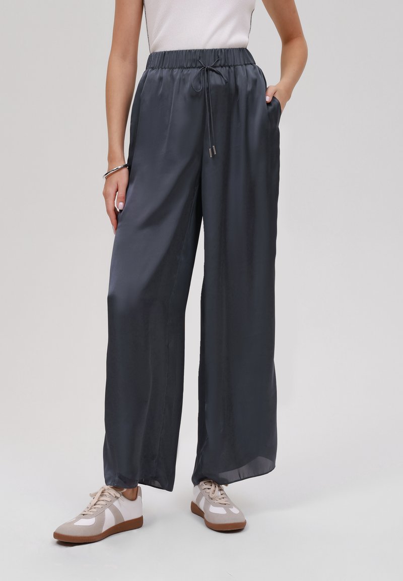 Pantalons larges en satin léger gris foncé avec une taille élastique, un cordon de serrage et des poches latérales, portés avec des baskets beiges.