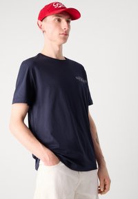 Marineblaue Baumwoll-T-Shirt mit kurzen Ärmeln und Rundhalsausschnitt, mit einem kleinen Logo auf der Brust. Getragen mit hellen Hosen.
