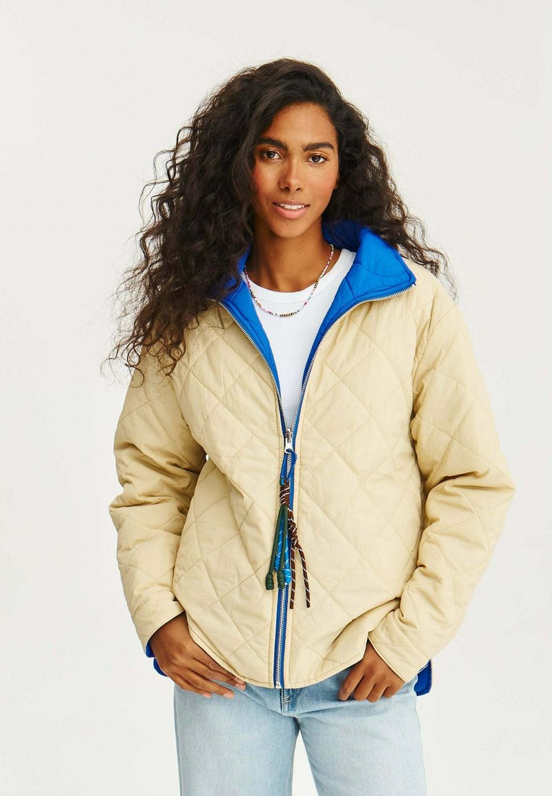 Béžová prošívaná bunda s modrým límcem a zipem, s úzkým pasem a barevnými táhly zipu, nošená přes bílý top a džíny.