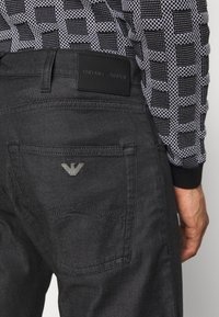 Svarta jeans med en slät yta och en silverfärgad örnlogga på bakfickan; har en läderetikett från Emporio Armani på midjebandet.