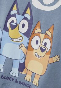 Plava pamučna majica s crtežima pasa, jedan plavi s žutim naglaskom i jedan narančasti. Ispod piše "BLUEY & BINGO" krupnim slovima.
