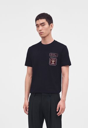 Jovem usando uma t-shirt preta com dois patches bordados cor de vinho no peito e calças escuras plissadas.
