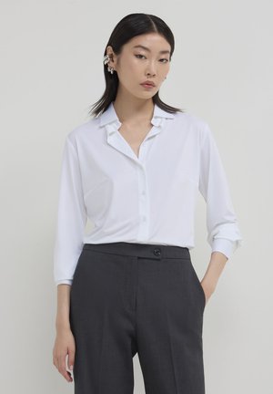 Donna con capelli scuri e lisci, indossa una camicia bianca con colletto e maniche arrotolate e pantaloni alti grigio scuro, in piedi davanti a uno sfondo semplice.