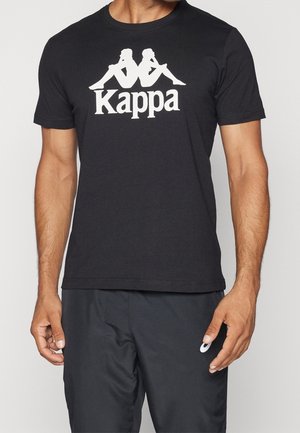 Mężczyzna ubrany w czarny t-shirt Kappa z białym logo przedstawiającym dwie sylwetki siedzące plecami do siebie oraz czarne spodnie z małym białym logo.