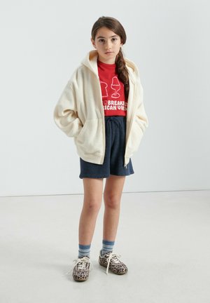 Kremna zip-up jopa iz flisa, rdeča majica z motivom, mornarsko modre kratke hlače, črtaste nogavice in rjavi čevlji z leopardjim vzorcem. Enostavna podlaga.