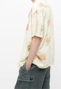Camicia beige a maniche corte con grafiche a tema tropicale in arancione e verde. Vestibilità rilassata, tessuto leggero, aperture laterali per maggiore comfort.