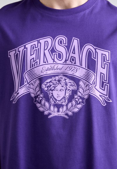 Lila póló nagy "VERSACE" felirattal, "Alapítva 1978" alcímmel, és laurel levelekkel keretezett Medúza fej logóval az elején.